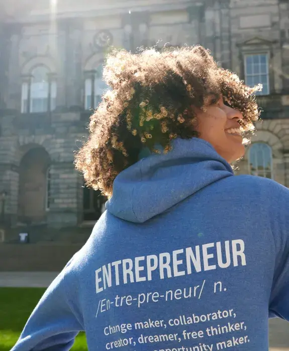 Student wearing a sweatshirt saying "entrepreneur" stands in front of Old College Edinburgh