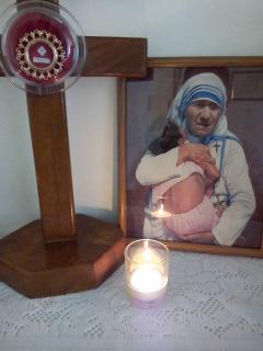 Photograph of a cross, a lit candle and a photoframe of Mother Theresa holding a child. 