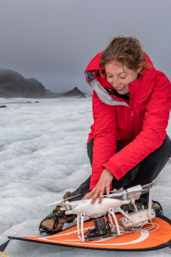 A female scientist in snow gear on a glacier kneeling down and holding a flying drone in her hands