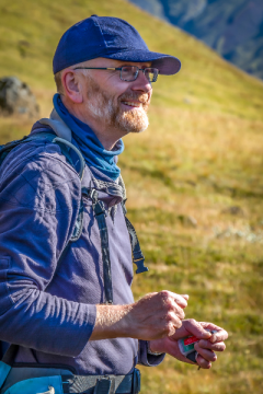 A male academic wearing a hat and weather gear smiling as he stands and teaches on a hillside in Iceland