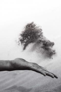 Black and white image of a hand outstretched. Above the hand is grains of sand that are flying after being thrown. 