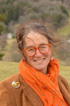 Photograph of Lesley Morrison. Lesley is wearing an orange scarf and a brown jacket. She is standing outside in front of some tr