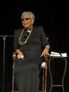Photograph of Maya Angelou, sitting in a chair on a stage in front of a microphone. 