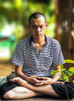 Photograph of a man sitting outside meditating. He is sitting with his legs crossed, his eyes closed and his hands in his lap. In the background there is trees and green plants around him. 