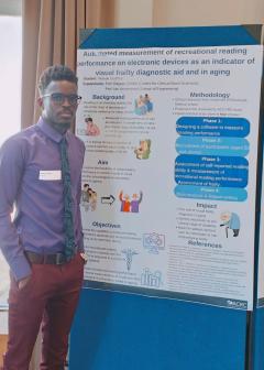 Wanok stands in a blue shirt in front of a posterboard. The poster is the title of his thesis.