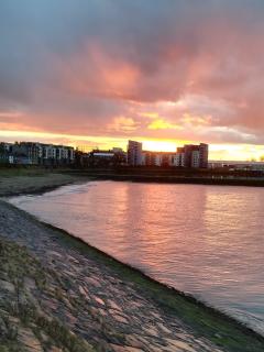 Photograph of the calm water at Wardie Bay, in the background the sun is setting behind some tall buildings. 