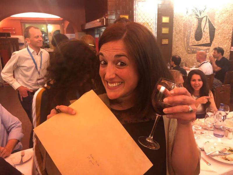 Alba, sitting at a table with her prize envelope, raises a glass