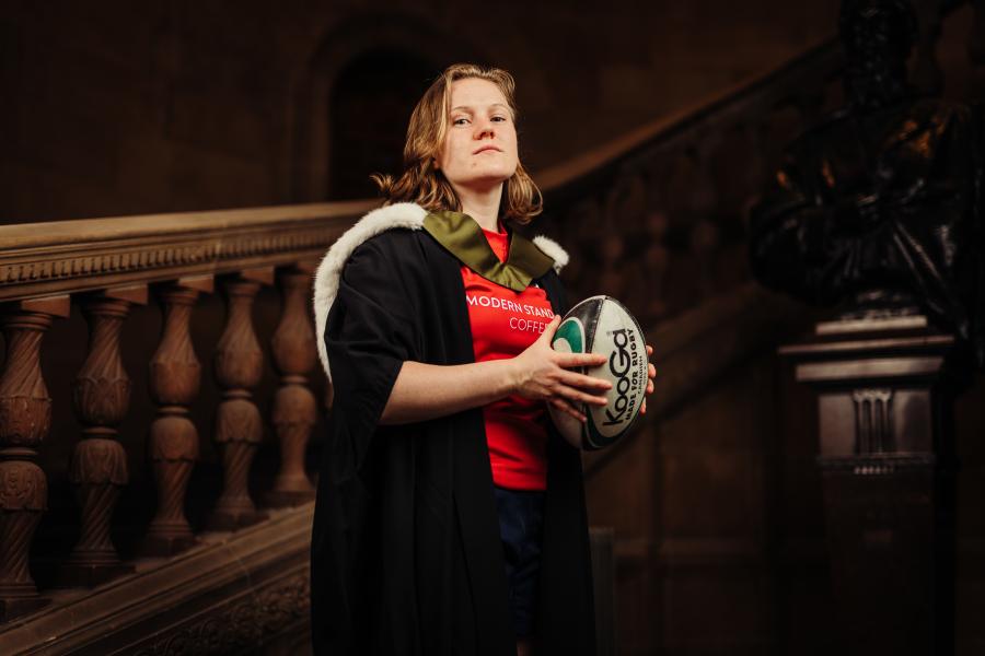 Meryl Smith wearing graduate gown and sports kit holding a rugby ball by McEwan Hall staircase