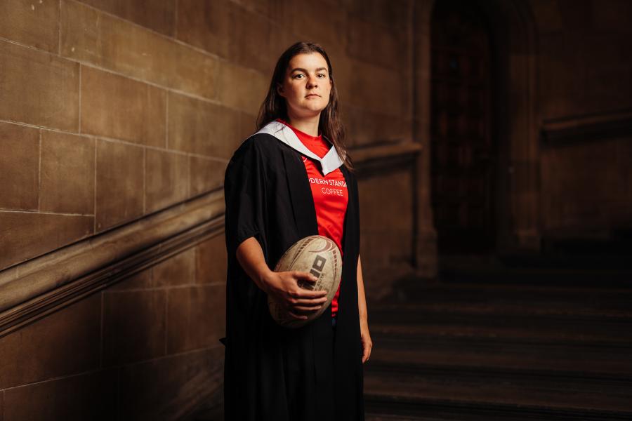 Sarah Denholm wearing graduate gown and sports kit holding a rugby ball on McEwan Hall staircase