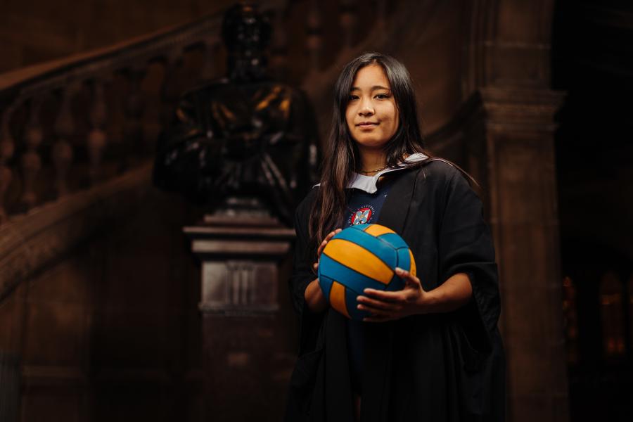 Caitlin Dila wearing graduate gown and sports kit holding water polo ball by McEwan Hall staircase