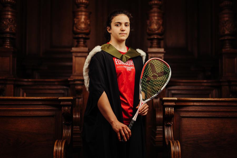 Georgia Adderley wearing graduate gown and sports kit holding a squash racket in McEwan Hall 