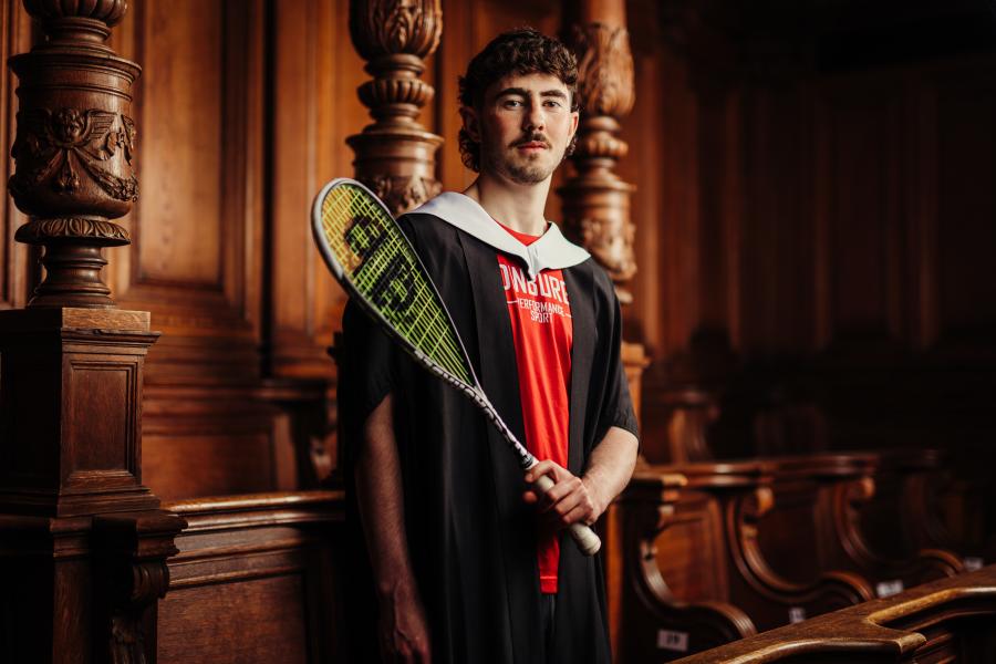 John Meehan wearing graduate gown and sports kit holding a squash racket in McEwan Hall 