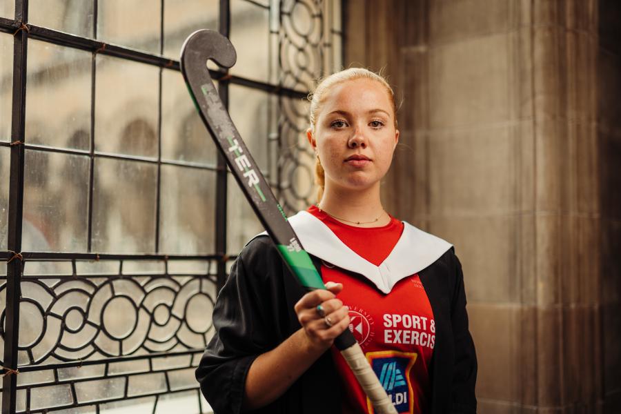 Eilidh Campbell wearing graduate gown and sport kit holding hockey stick in McEwan Hall by stained glass windows