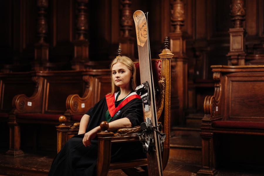 Emma Olley wearing graduate gown and sports kit sitting on red leather chair in McEwan Hall with skis propped against chair