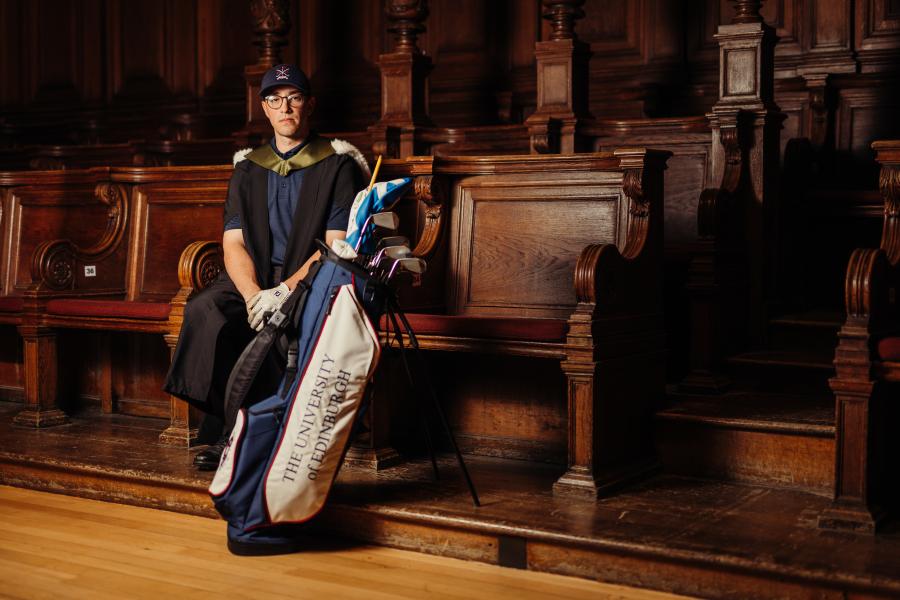 Max Holford wearing graduate gown and sports kit sitting on stage benches in McEwan Hall with golf bag in front